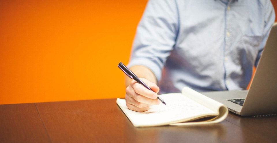 imagem de um homem sentado na mesa de trabalho escrevendo ao lado do notebook trabalhando em uma tradução juramentada basileia